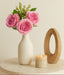 White vase with pink roses on a round wooden table with a candle and wooden ring.