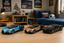 Three toy sports cars resembling a Bugatti Chiron are lined up on a light wood tabletop in a bedroom setting. The cars are in metallic blue, metallic gold, and metallic black with yellow accents.