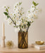 Decorative setup with white cherry blossom flowers in a vase, candle, and small bowl on a wooden surface.