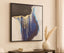Abstract art piece with gold and blue elements on a beige wall, accompanied by a vase and books.