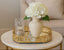 Decorative table setting with a vase of white flowers on a gold oval mirror tray.