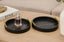 Two black marble textured trays with gold handles on a wooden surface, one holding a perfume bottle.