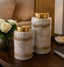 Two marble-patterned jars with gold lids on a wooden surface, with flowers and a bowl in the background.