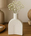 White ceramic vase with arches holding small white flowers on a wooden surface.