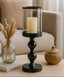 Decorative candle holder with a candle on a table next to a vase and small jar.