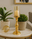 Gold candle holder with a lit candle on a marble table with plants and a gold decorative object.