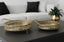 Two gold decorative mirror trays on a dark surface with a white sofa and plant in the background.