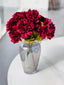Two maroon artificial bloomed peony bunches styled in a smoked glass vase on a marble table.