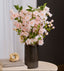 Vase with pink cherry blossom flowers on a table against a white curtain and dark wall