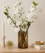 Decorative setup with white cherry blossom flowers in a vase, candle, and small bowl on a wooden surface.