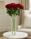 Six red artificial rose stems in a clear glass vase on a round white table beside a sofa.