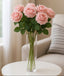 Six soft pink artificial rose stems in a clear glass vase on a round white table.