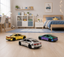 Three miniature toy cars with metallic finishes are displayed on a light wood floor in a modern, well-lit children's bedroom.