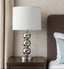 Silver Table lamp with a white shade on a wooden nightstand next to a bed.