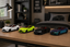 Four miniature toy sports cars in white, neon yellow, black, and blue-purple are lined up on a wooden desk in a modern bedroom setting.