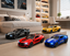 Four toy sports cars in black, red, yellow, and blue are arranged on a light wood floor in a modern bedroom setting. The cars have 'LBWK' printed on the sides and are styled as if they are real cars, with a bed, shelves, and desk visible in the background.