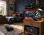 A toy truck box labeled 'Rock Crazzy' sits on a wooden nightstand in a styled children's bedroom. The room features a blue wall with climbing decals, a bed with blue bedding, and a toy truck on the rug nearby.
