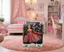 A boxed fashion doll with a long blonde dress and matching accessories stands on a round pink rug in a pink-themed bedroom. The room includes a bed, shelves, desk, chair, and string lights.