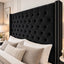 A large black tufted velvet headboard with silver nailhead trim, shown in a styled bedroom setting with neutral beige and white bedding.