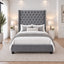 A grey upholstered bed frame with a tall tufted headboard and matching footboard, styled in a bright, modern bedroom with white bedding, striped pillows, bedside tables, lamps, and a large area rug.