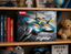 Box packaging for a DHM 2.4CHZ Fighter drone toy, displayed on a wooden shelf next to books, a teddy bear, and other toys.