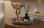 Decorative items including a gold champagne glass with a pink rose, a black diffuser bottle on a grey tray and books on a wooden table.