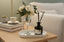 Decorative setup with flowers in a champagne glass, and a diffuser on a table.