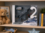 Boxed RC combat aircraft set displayed on a wooden shelf, featuring a gray fighter jet toy, remote control, and printed packaging with the text 'CROUDNX' and 'Combat Aircraft Fighter'. Surrounding props include a teddy bear, books, a small airplane, and a star-shaped decor piece.