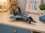A gray toy model of a fighter jet displayed on a light wood desk in a child's room setting, surrounded by a teddy bear, a lamp, a framed sign, and a small potted plant.