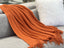 Cozy Levi Throw Rug Blanket in orange draped over a grey sofa with white cushion, showcasing soft texture and fringe.