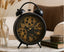 Decorative black and gold alarm clock on a table with books and a vase in the background