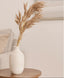 White vase with dried pampas grass on a wooden side table against a neutral wall.