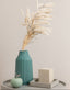Teal vase with pampas plants on a gray surface against a light gray background