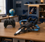 A black remote-controlled toy airplane with blue and white wing accents is displayed on a wooden table next to its black controller in a styled room setting.