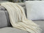 Cream Tessa throw rug blanket draped over a grey sofa with a fluffy white cushion for a cozy touch.