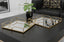 Two gold square mirror decorative trays on a dark surface with a plant and couch in the background.